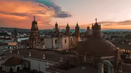 Aerial view of colonial Mexican church domes and bell towers silhouetted against a dramatic orange sunset sky.
