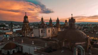 Aerial view of colonial Mexican church domes and bell towers silhouetted against a dramatic orange sunset sky.