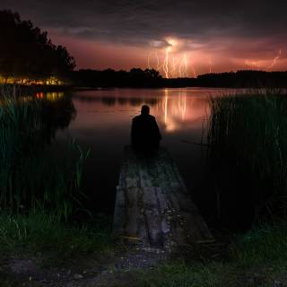 Silueta unui bărbat așezat pe un pod într-un iaz pe fundalul fulgerului care clipește pe cerul nopții