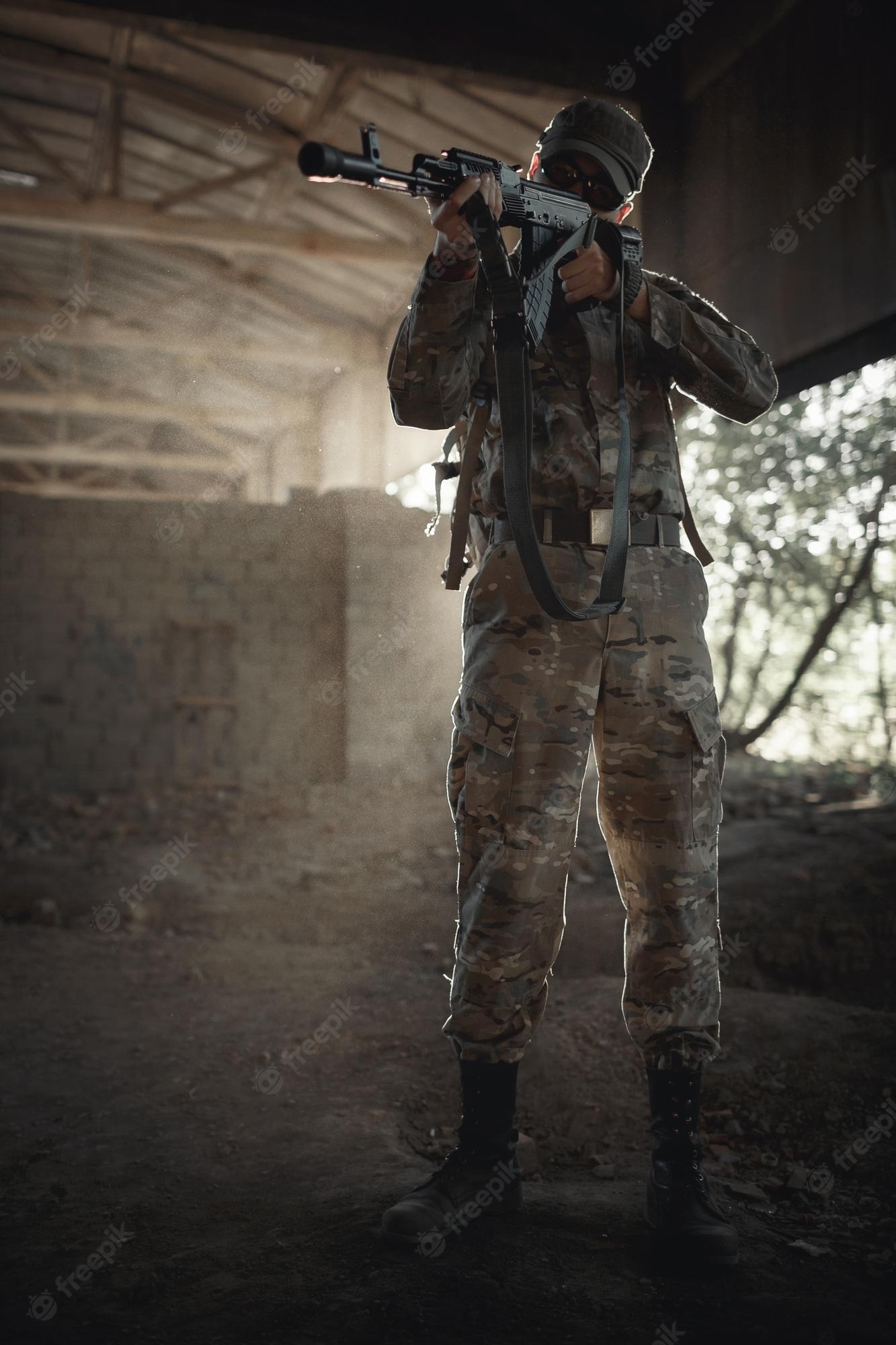 Premium Photo. A fearless soldier in a camouflage protective uniform in tactical black glasses and in a cap with a machine gun in his hands stands inside a large permitted house