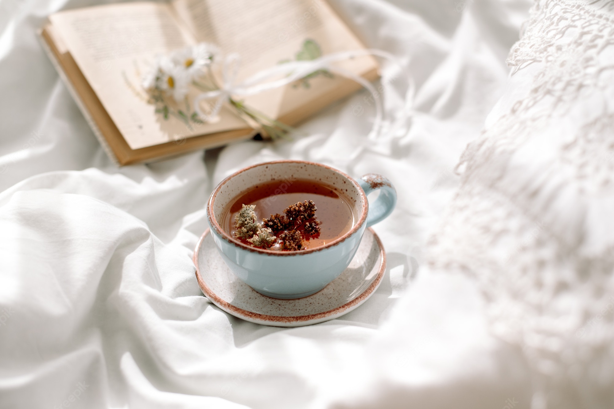 Premium Photo. Openwork lace, cotton white blanket. book botany, mug with natural herbal tea made from mint, summer daisy flowers. morning breakfast in bed. provence retro style. clean cosiness and freshness