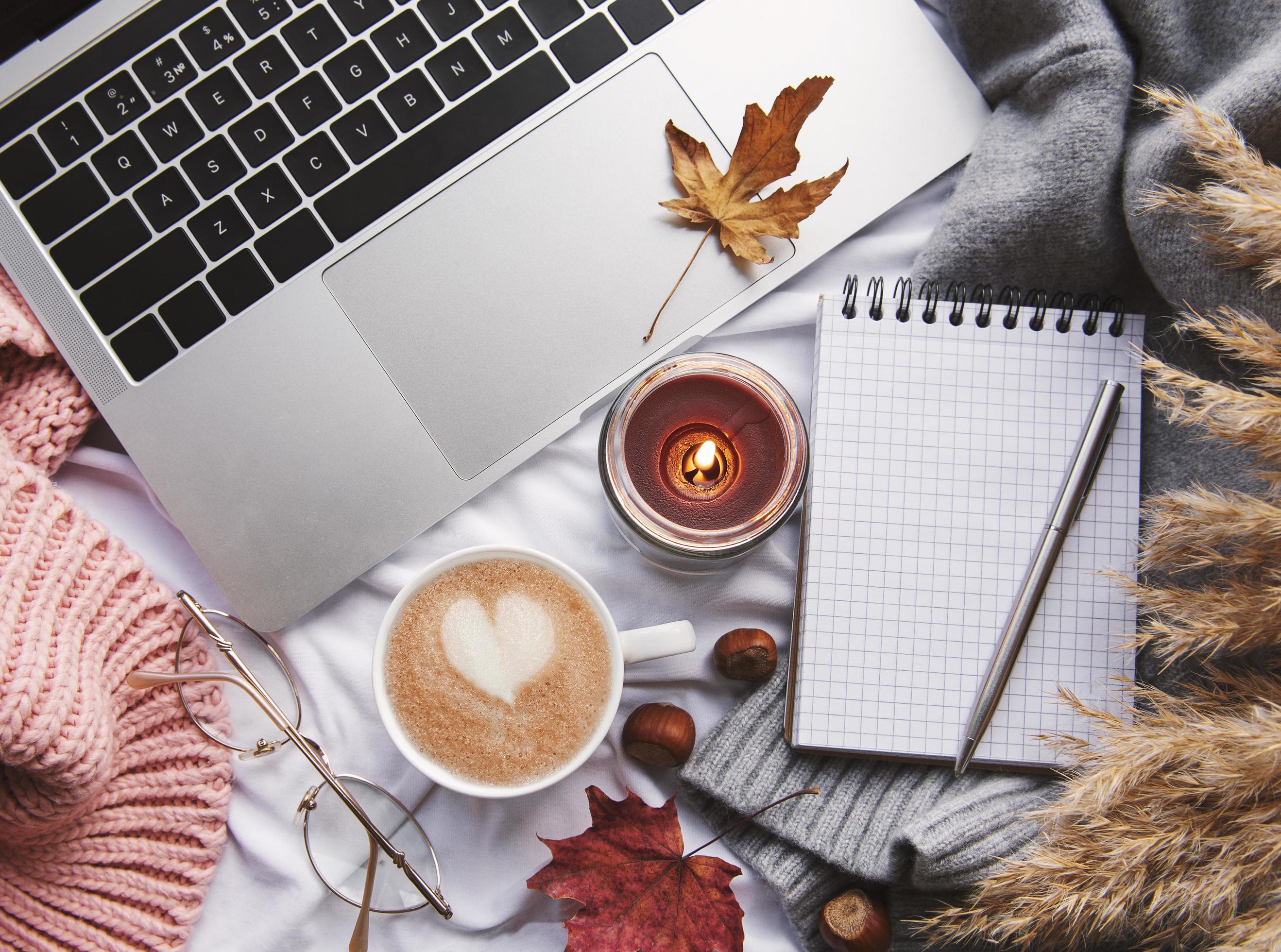 Laptop, sweater, yellow autumn leaves, candle and coffee on white bed