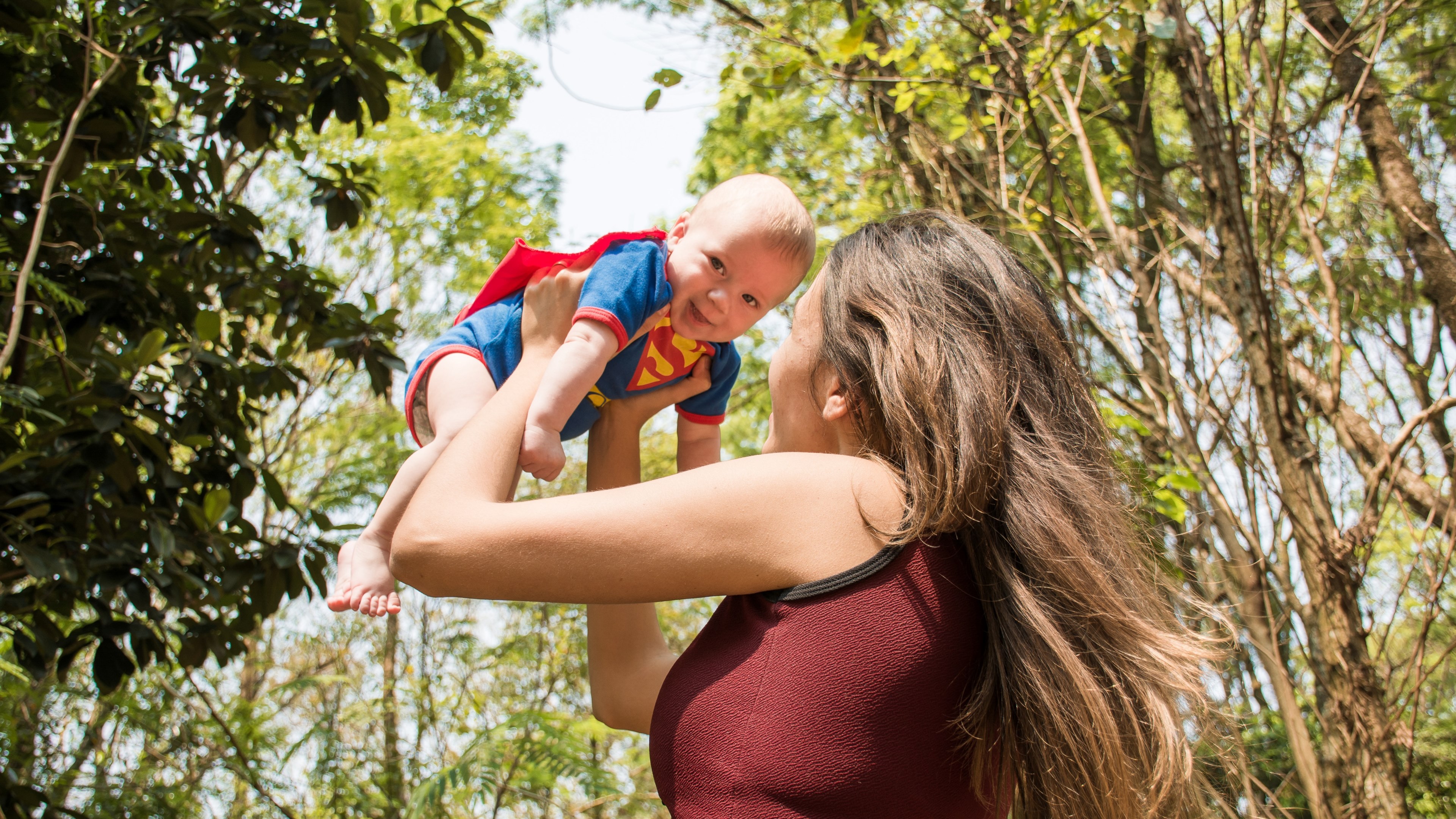 Wallpaper / a baby wearing a superman outfit being held in the air by his brunette mom in the forest at cianorte, superman baby flying in the forest 4k wallpaper
