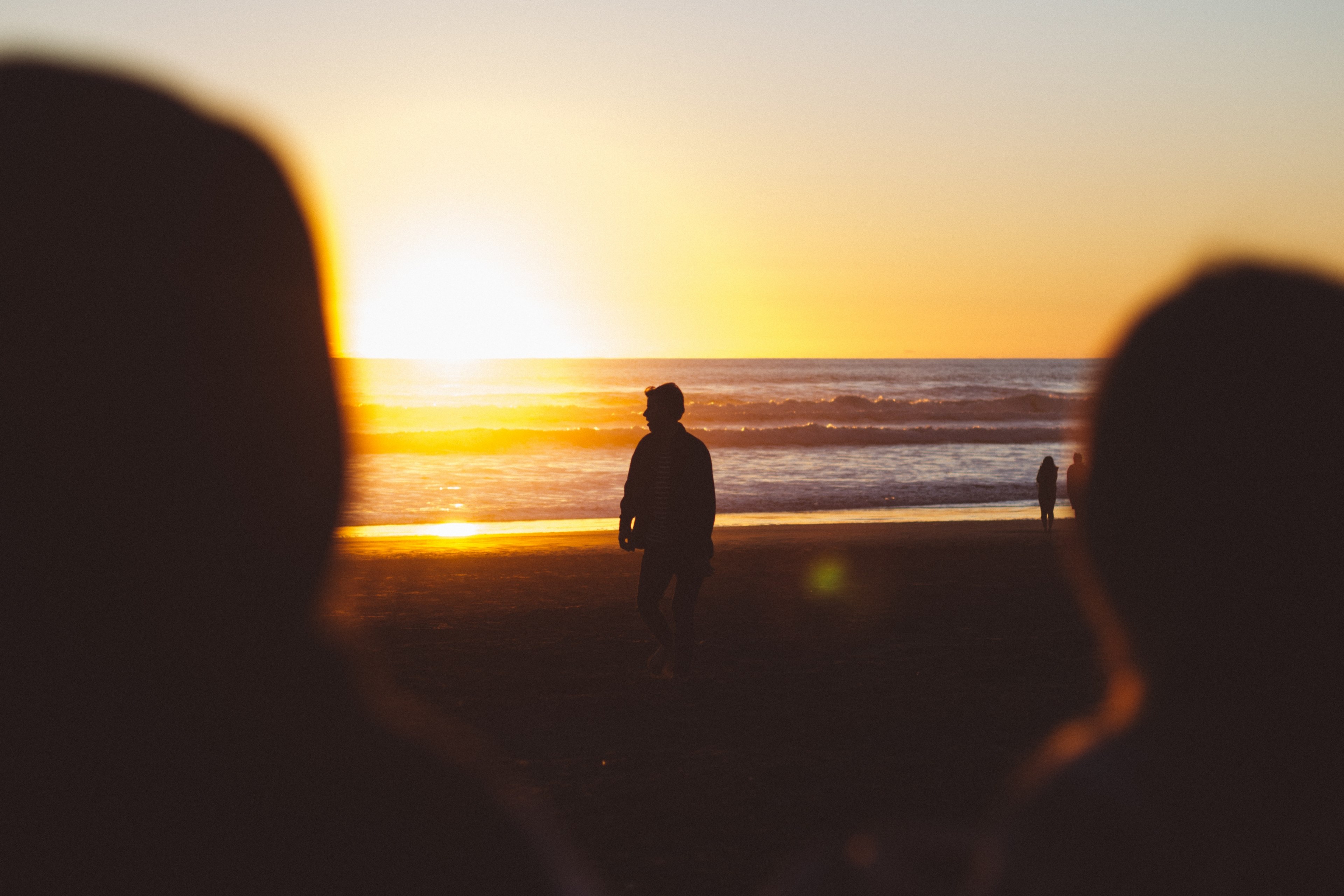 Wallpaper / silhouette of a person on the beach sun setting on the horizon seen over shoulders of two blurred people in the foreground, silhouette sunset sunrise and beach hd