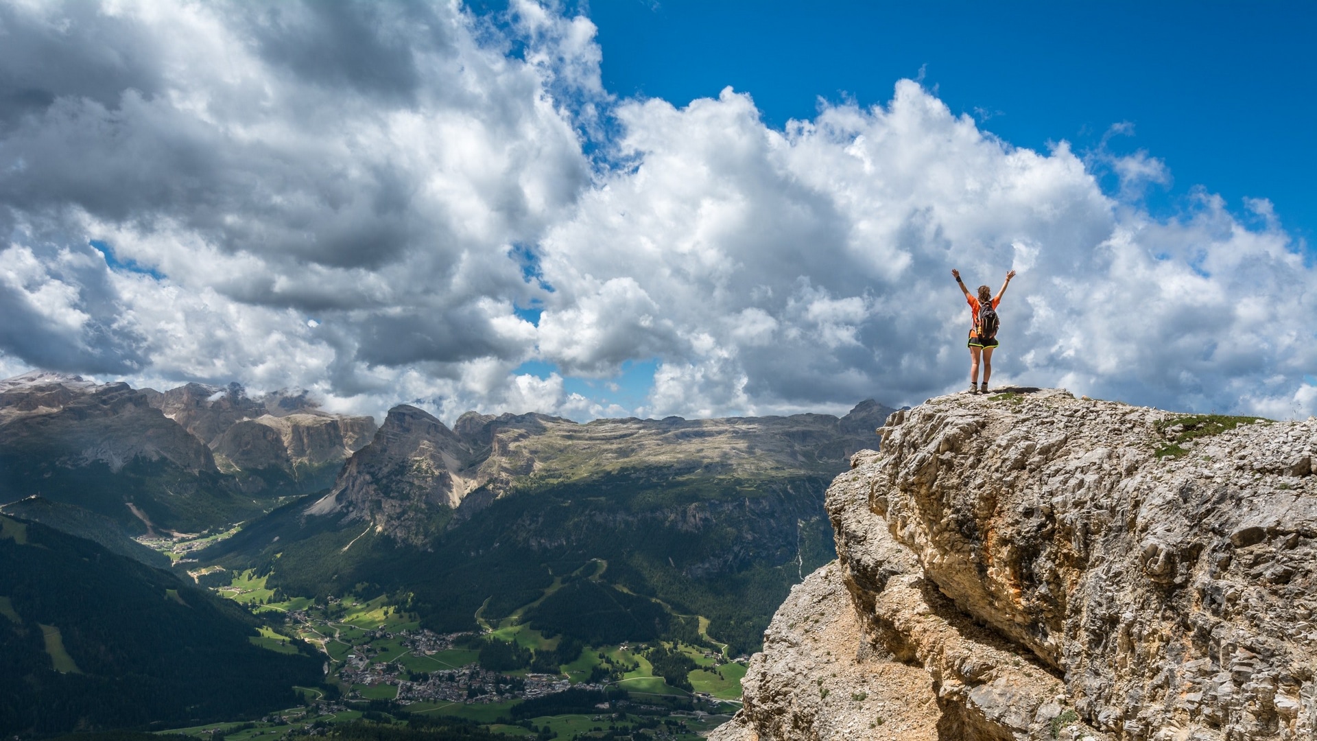 Wallpaper / a person throwing their hands up in the air on the edge of a tall jagged rock, looking down to the valley 4k wallpaper free download
