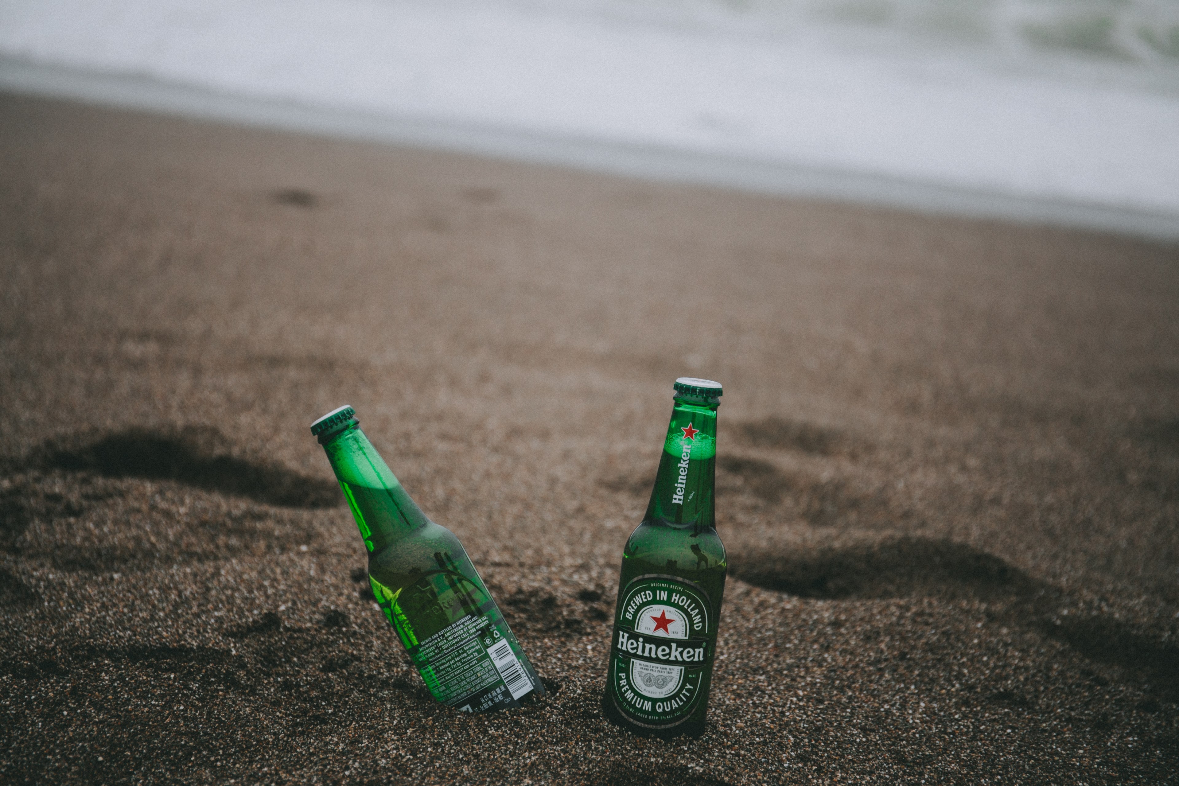 two heineken bottles beside each other one tilted placed on a sandy point reyes beach, beer by the beach 4k HD Wallpaper