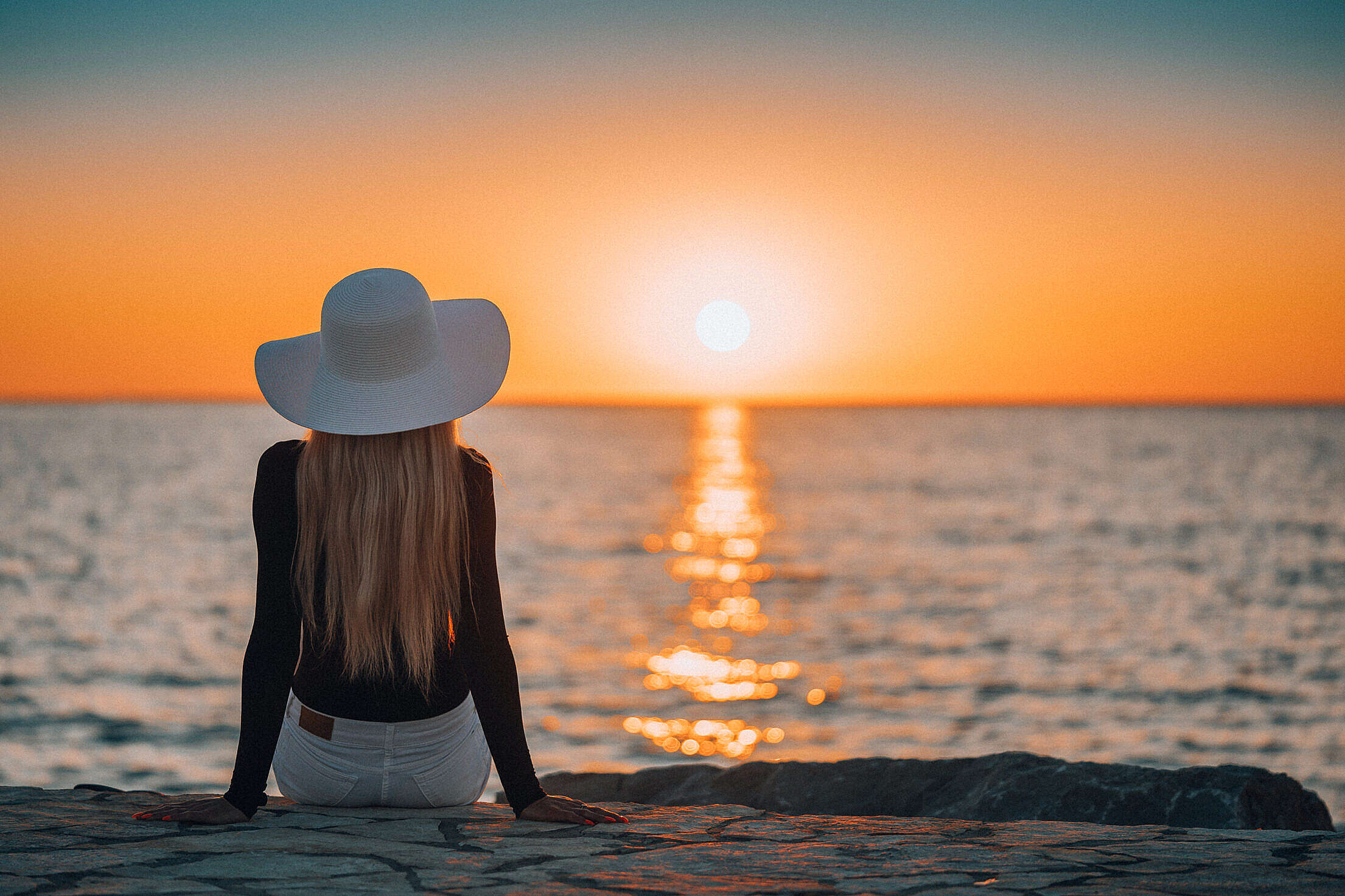Woman Sitting on a Rocky Shore by the Sea and Watching Sunset Free