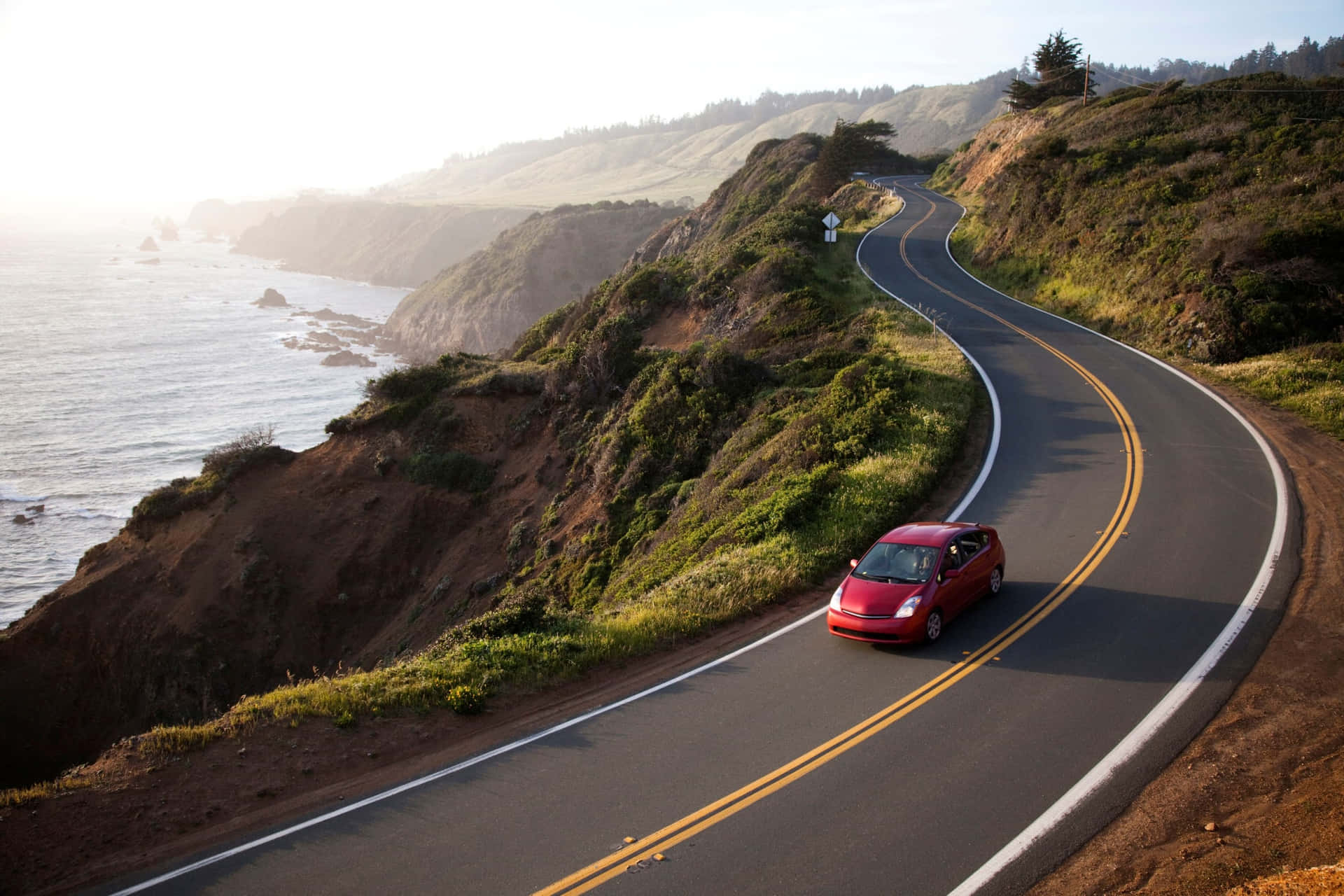 Download A Red Car Driving Down A Winding Road Near The Ocean