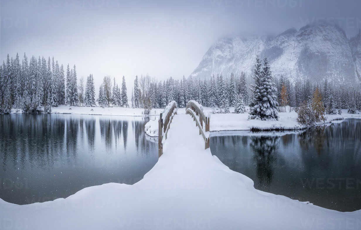 Beautiful winter Canadian landscape with snow covered bridge over calm water with fir forest and foggy snowy mountains in background