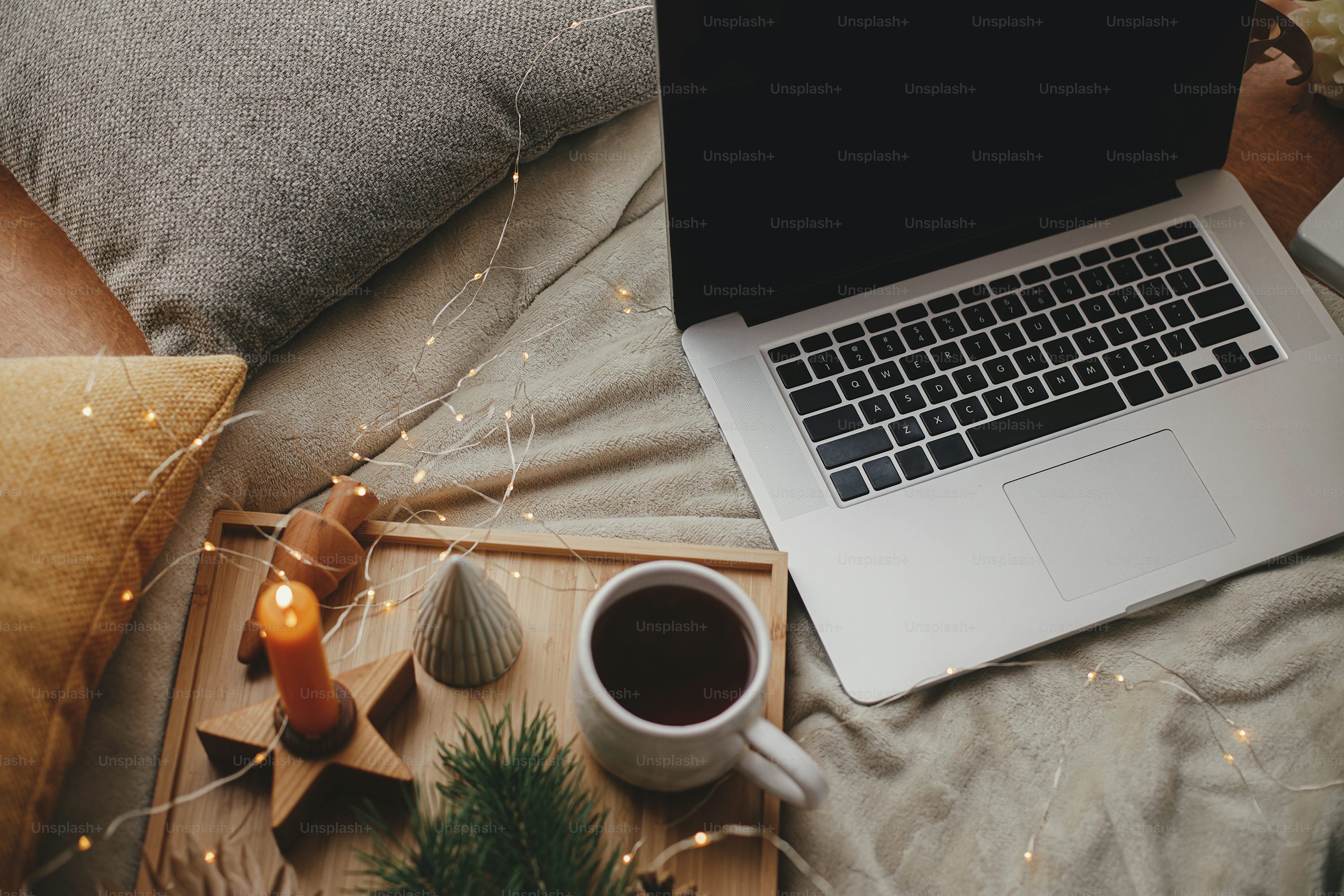 Laptop on soft bed with cup of tea, christmas lights, stars, pine, candle and pillows in scandinavian room. Freelance and blog. Remote work and online. Cozy winter holidays at home photo
