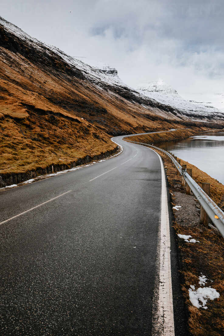 Spectacular view of wet winding road located near river and mountain slope with dry grass in winter on Faroe Islands