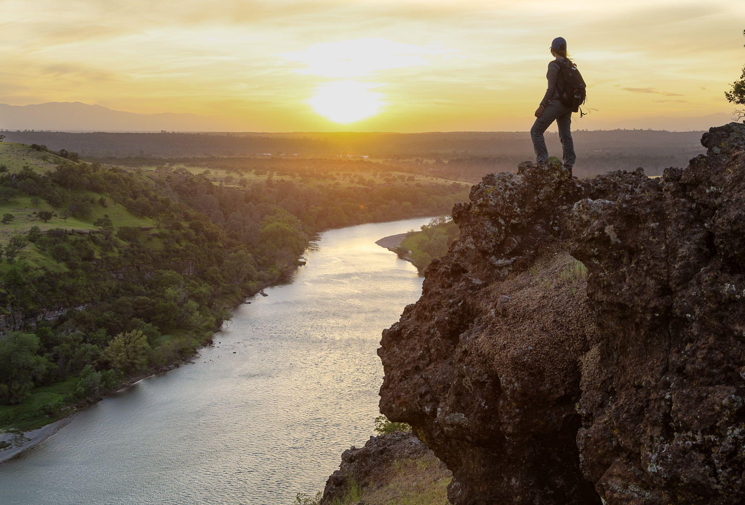 Man wearing backpack standing on rock cliff facing sunset infront