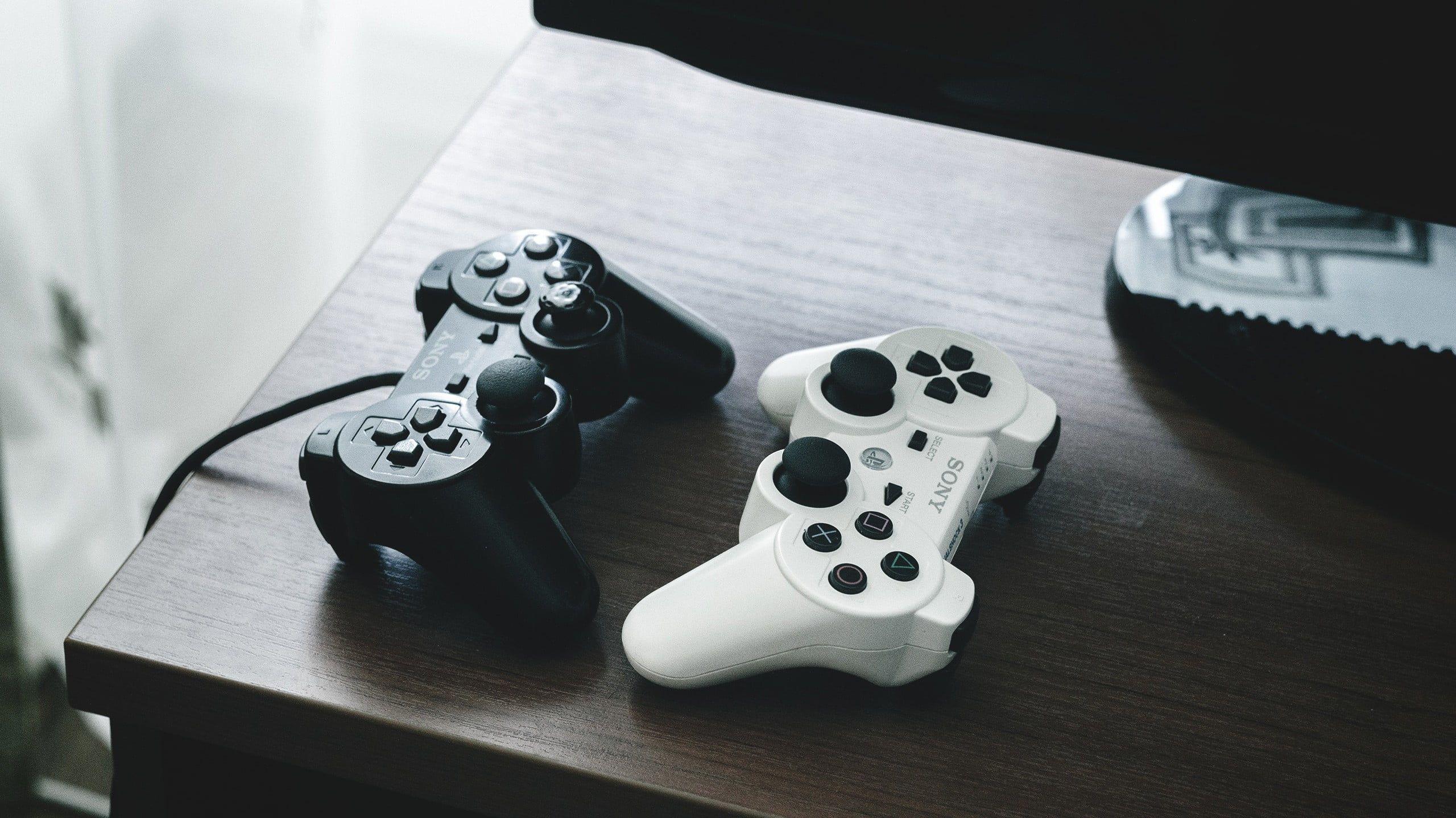 Two black and white Sony PS3 controllers on brown wooden table HD wallpaper