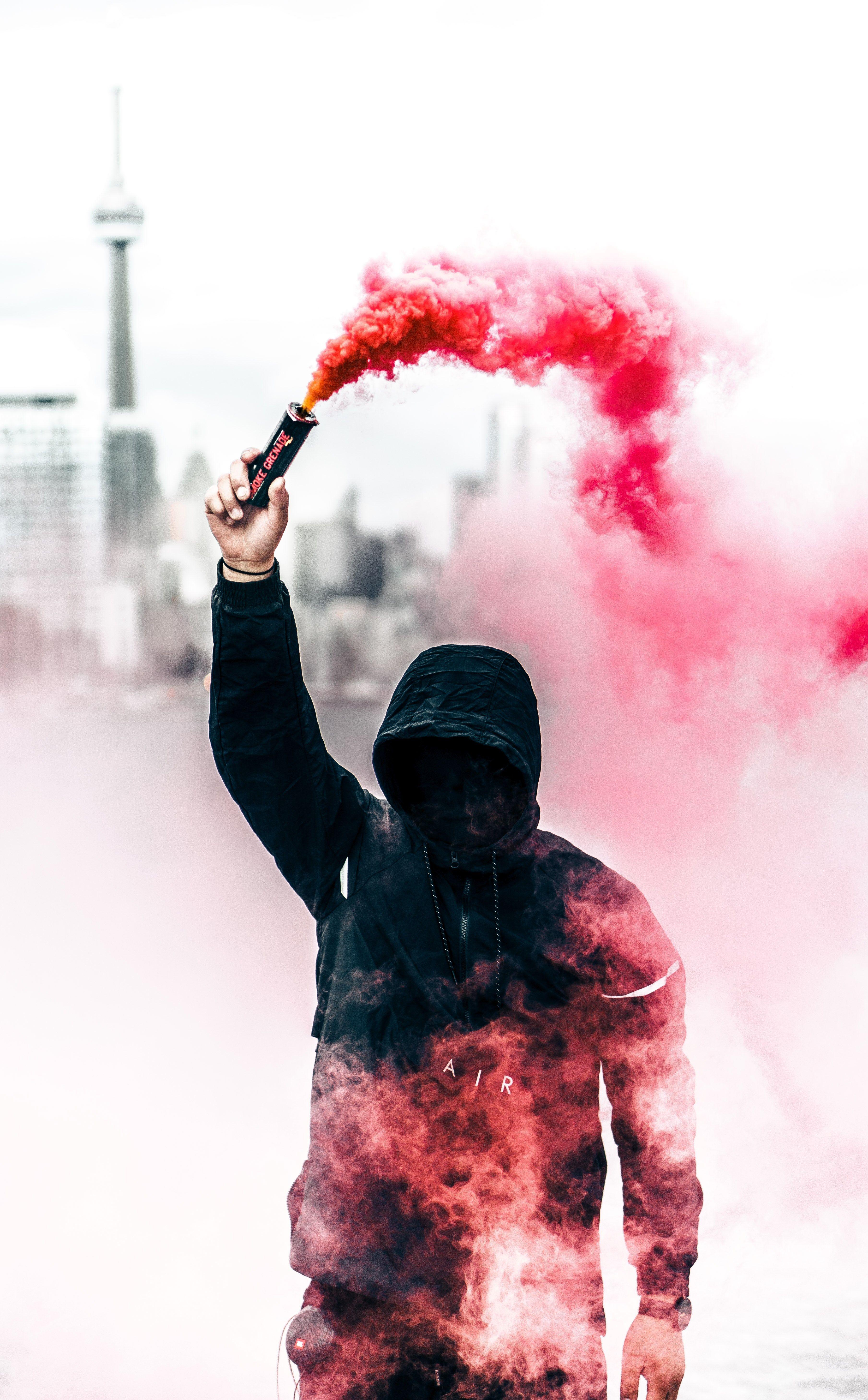 A person in a black hoodie with obscured face holds up a pink smoke