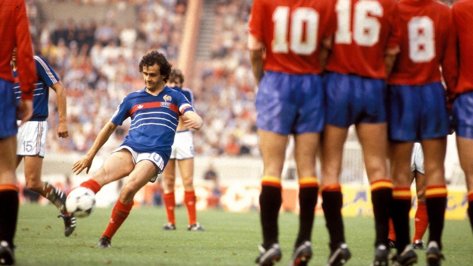  Michel Platini is seen scoring a goal against Spain in the Euro 84 tournament, wearing a blue jersey with white shorts and blue socks while the Spanish defenders wear red jerseys with blue shorts and red socks.