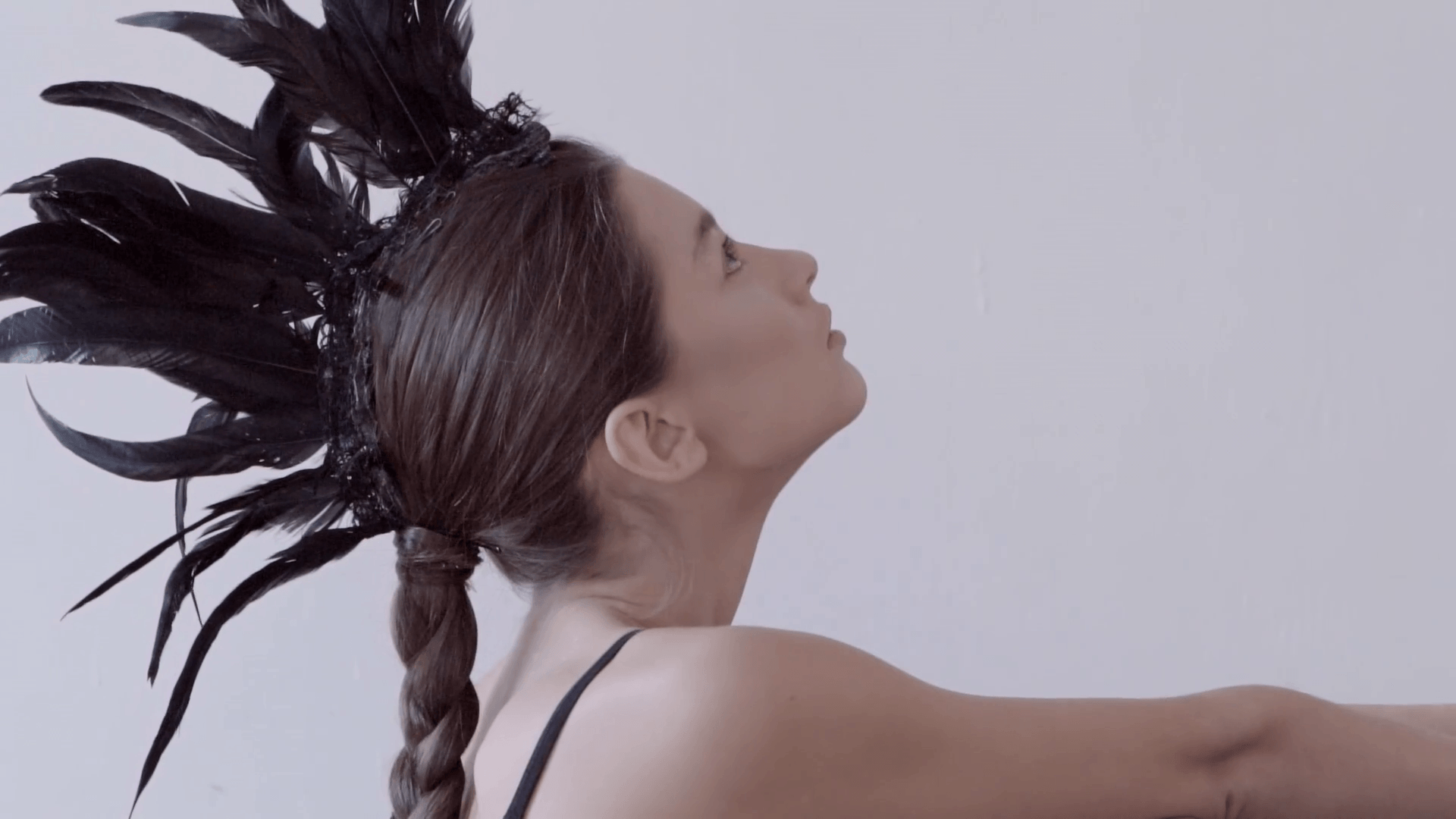 Young mixed race caucasian woman vogue portrait with feather mohawk accessory wearing black bodysuit sitting on wooden floor on white wall background. Fashion concept