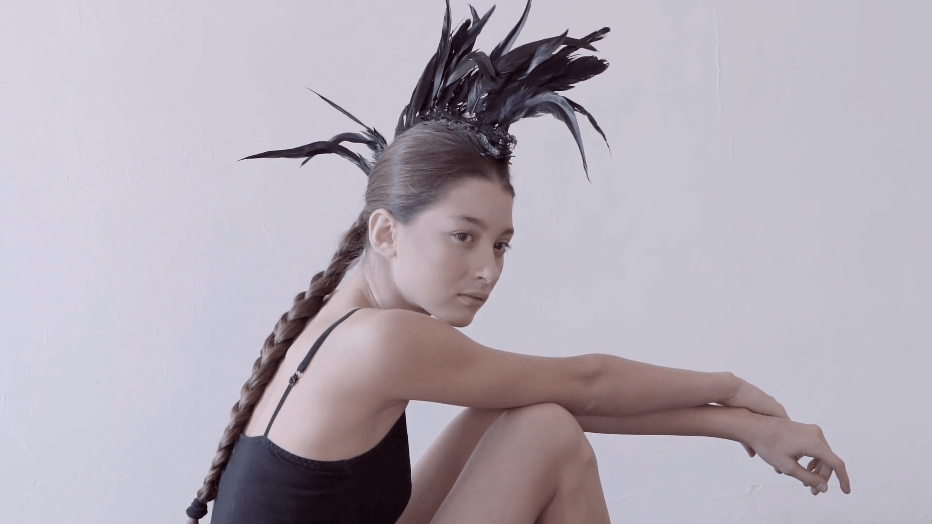 Young mixed race caucasian woman vogue portrait with feather mohawk accessory wearing black bodysuit sitting on wooden floor on white wall background. Fashion concept