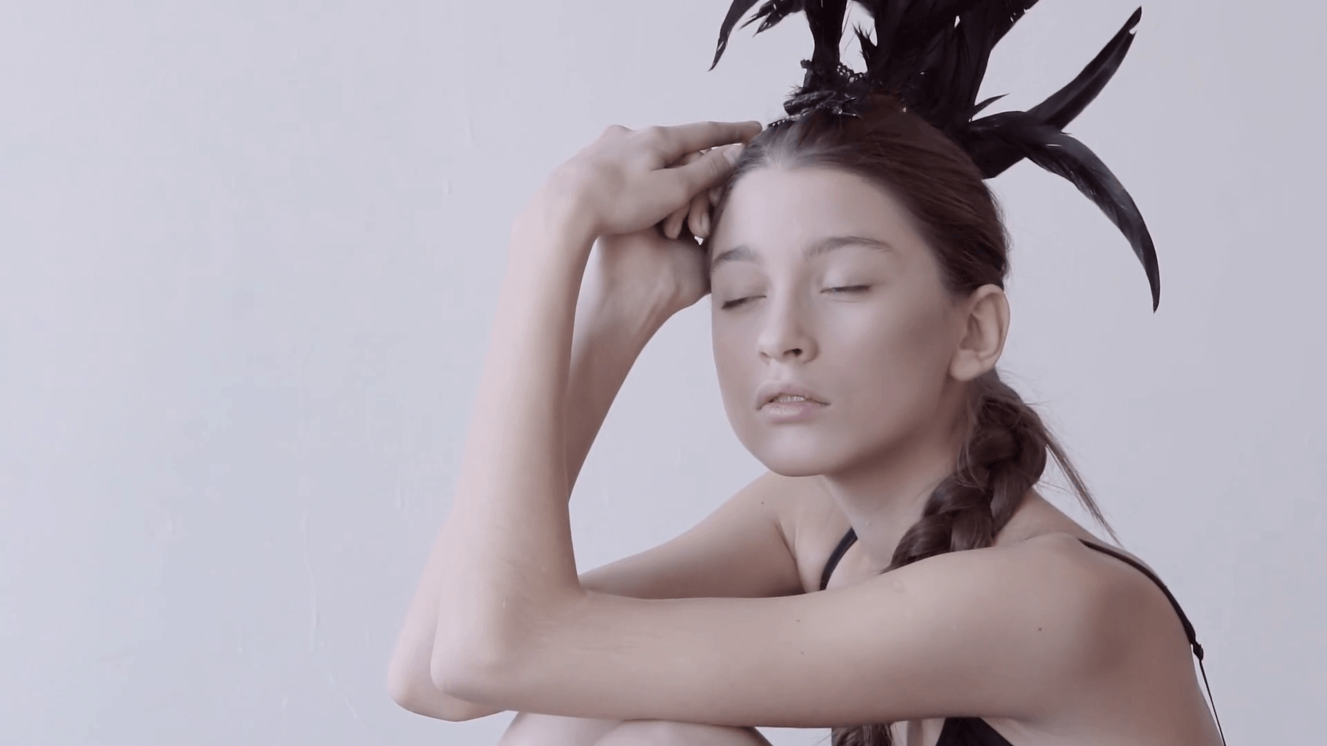 Young mixed race caucasian woman vogue portrait with feather mohawk accessory wearing black bodysuit sitting on wooden floor on white wall background