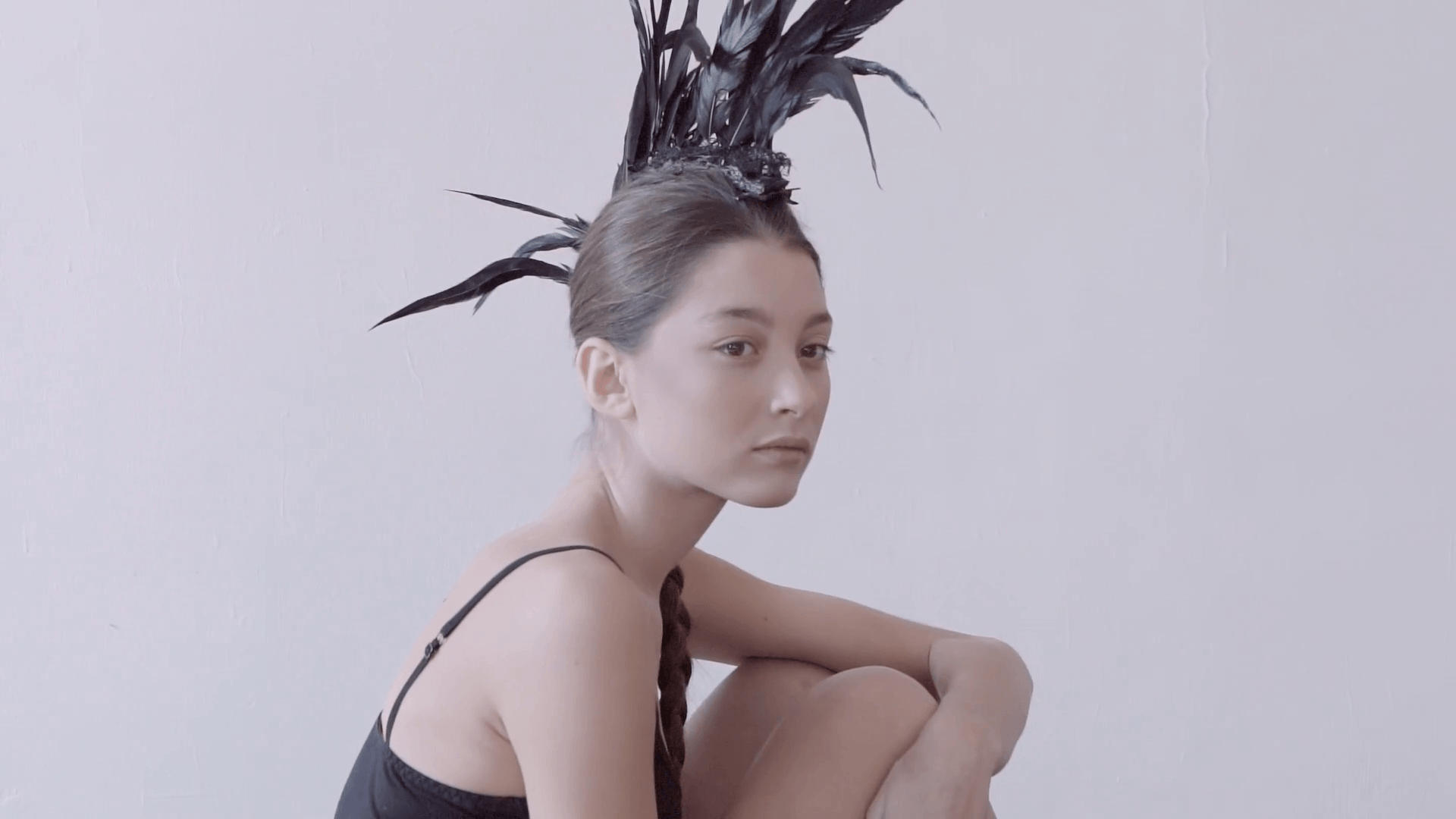 Young mixed race caucasian woman vogue portrait with feather mohawk accessory wearing black bodysuit sitting on wooden floor on white wall background. Fashion concept