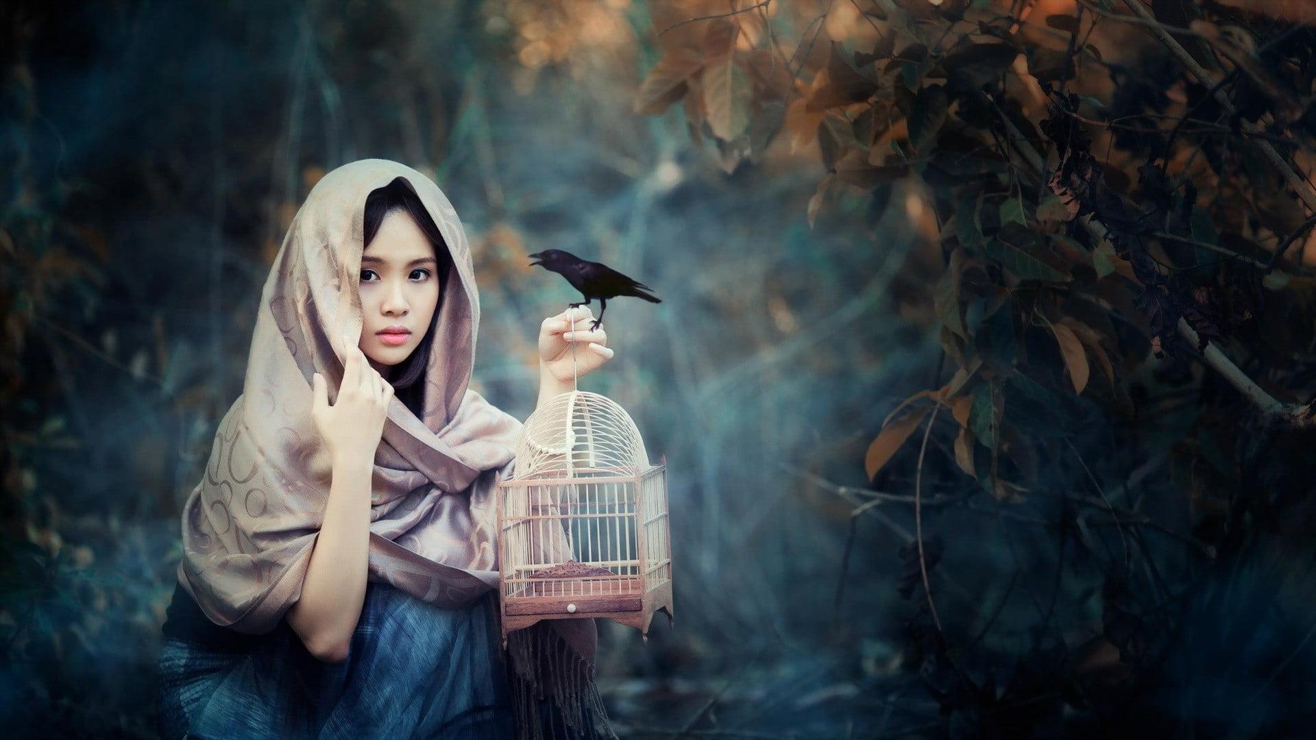 Woman wearing brown hijab holding black bird and brown bird cage