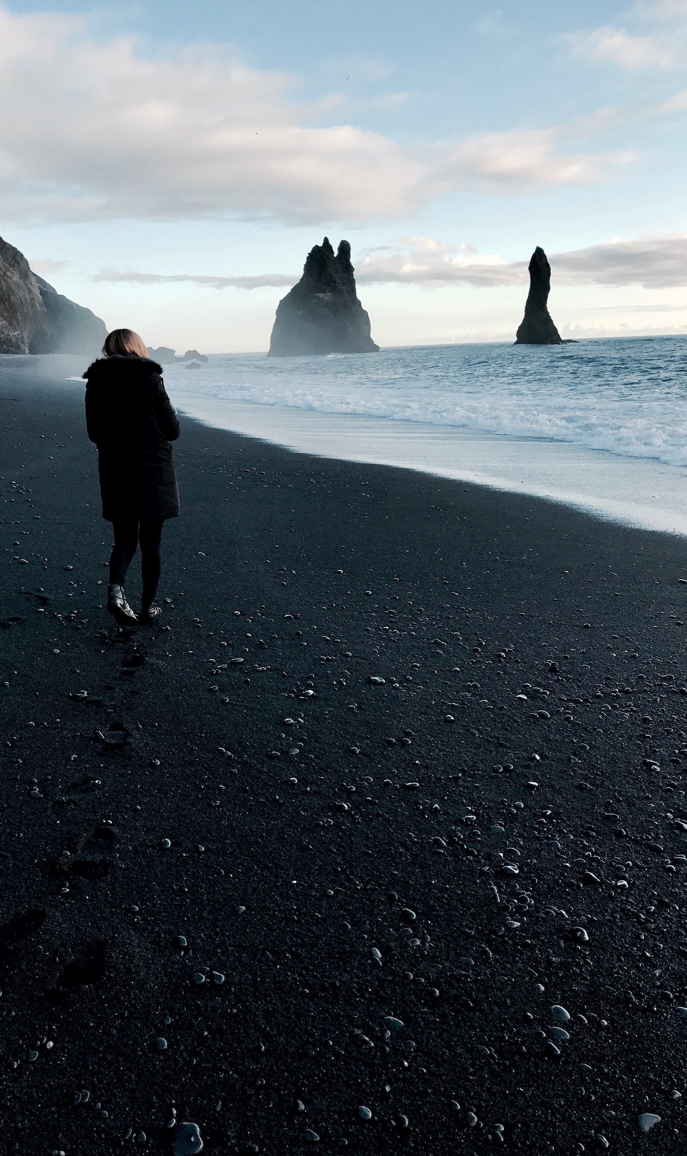 woman in black coat standing on a black sand beach looking