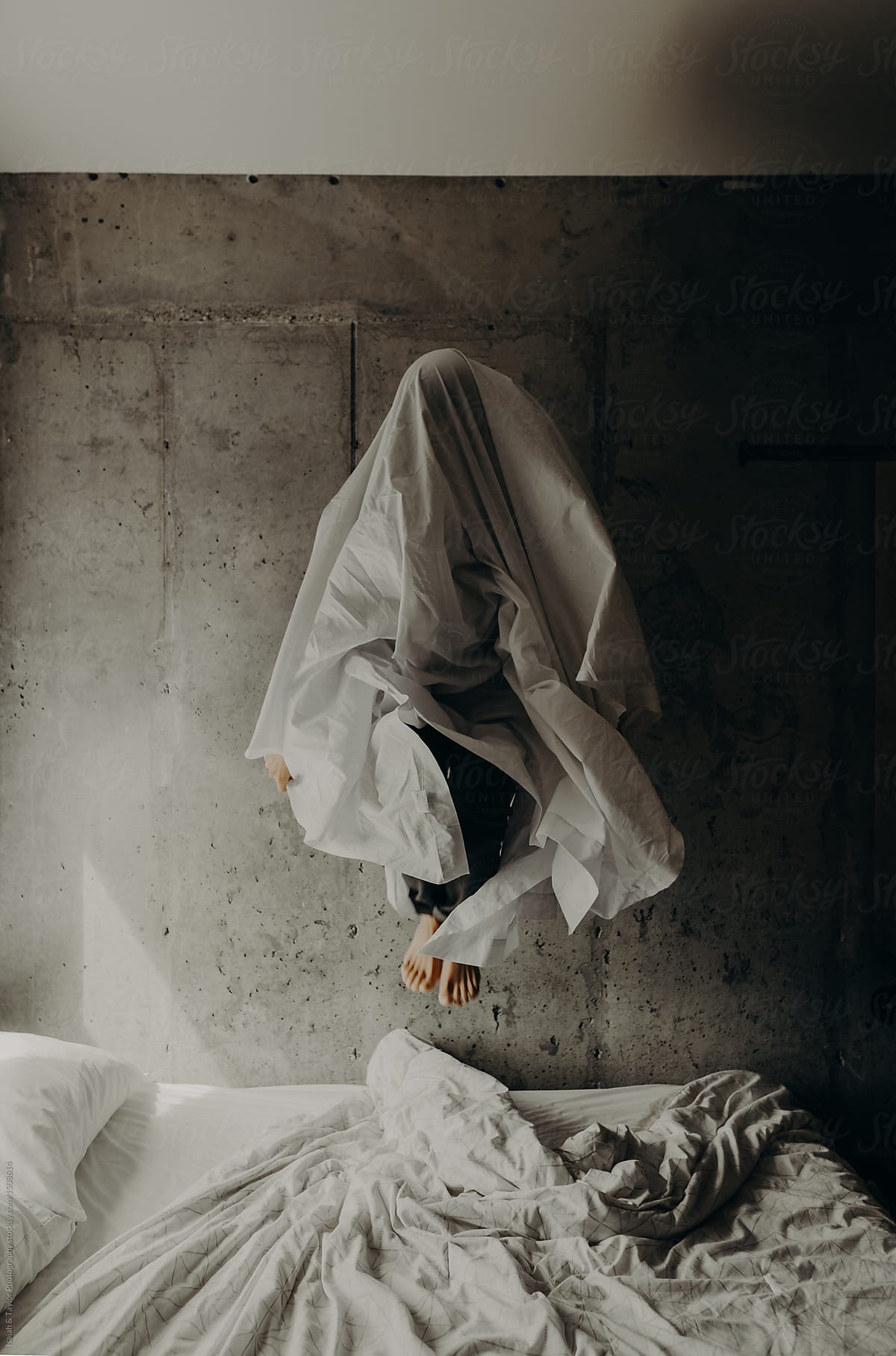 Ghost like figure of a woman wearing a bed sheet over her head while jumping on a bed by Isaiah & Taylor Photography
