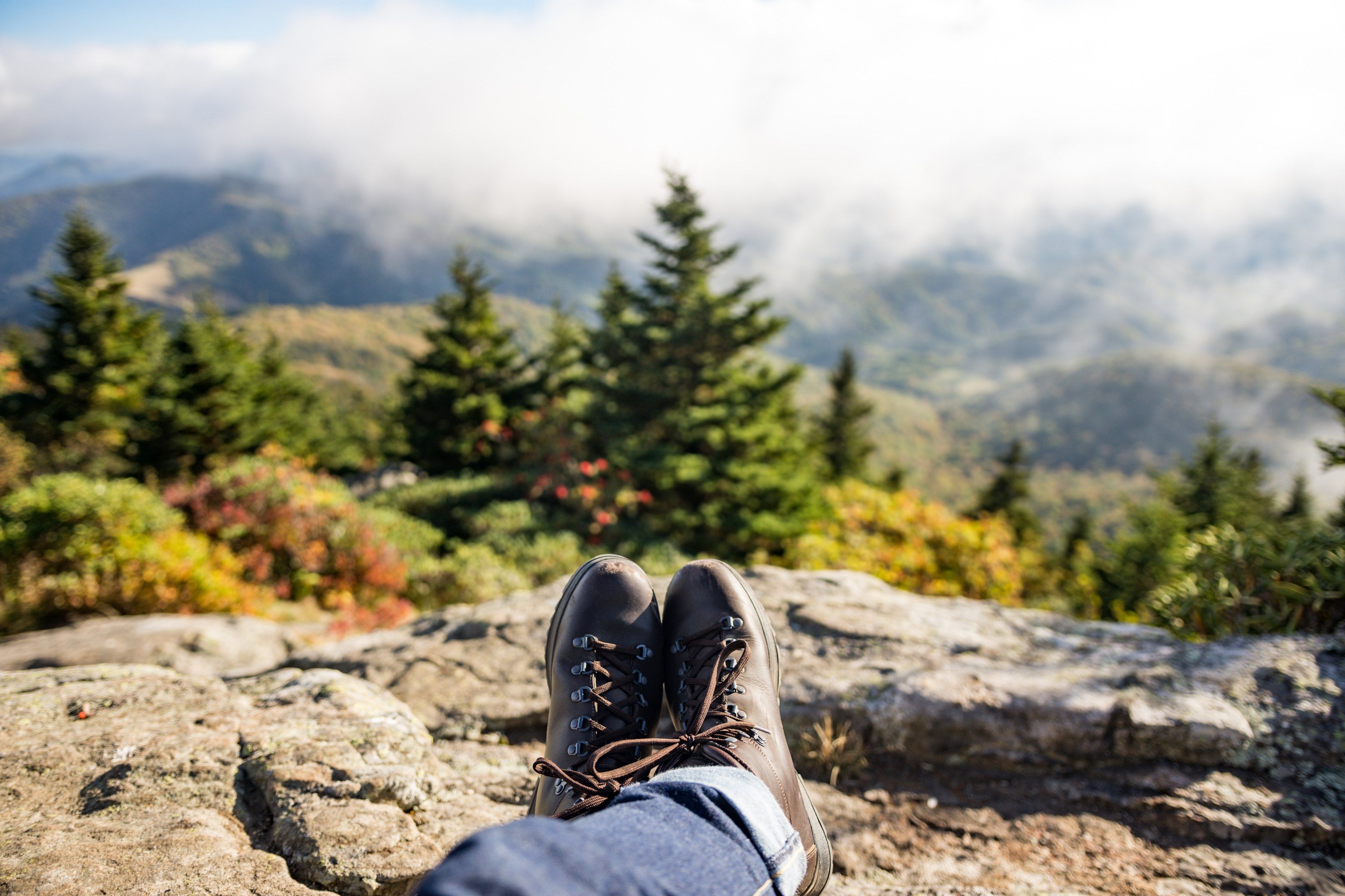 Wallpaper / a person wearing hiking boots and jeans resting on a rock looking out into the fog covered mountains, resting over a valley 4k wallpaper
