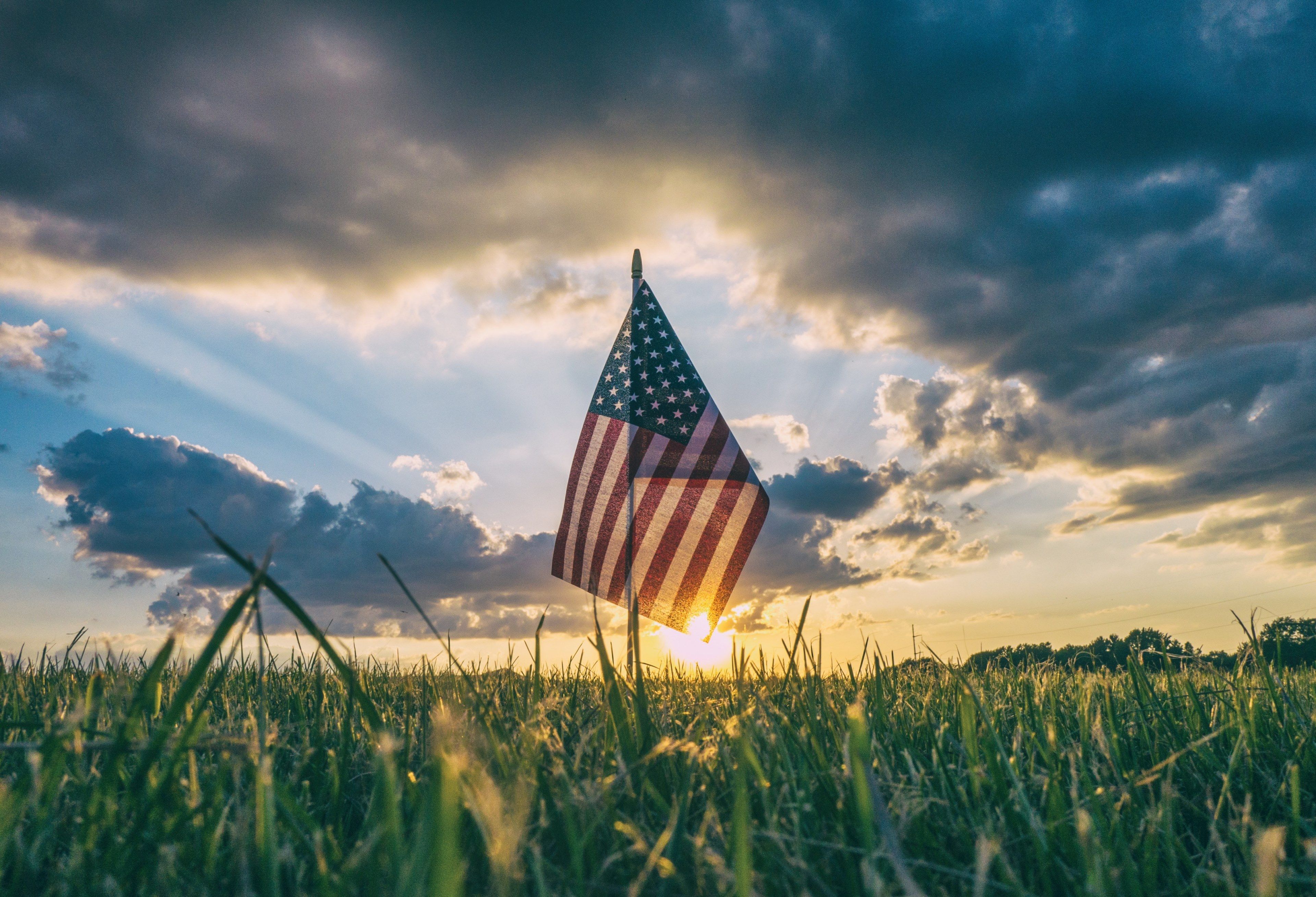 Wallpaper / the flag of america stands in the grass with looming sunnset on 4th of july, american flag in the grass 4k wallpaper
