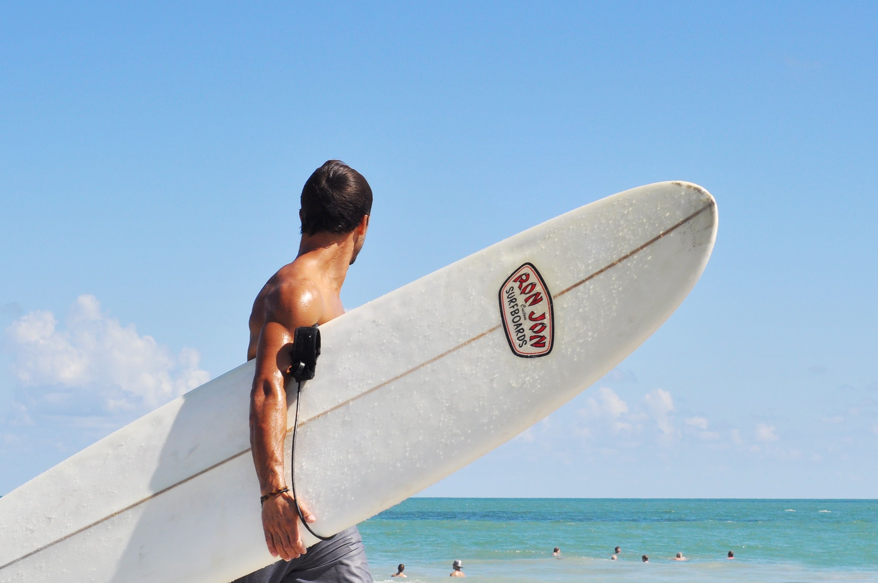 topless man carrying white ron jon surfboard and looking towards people swimming on beach under blue sky free image