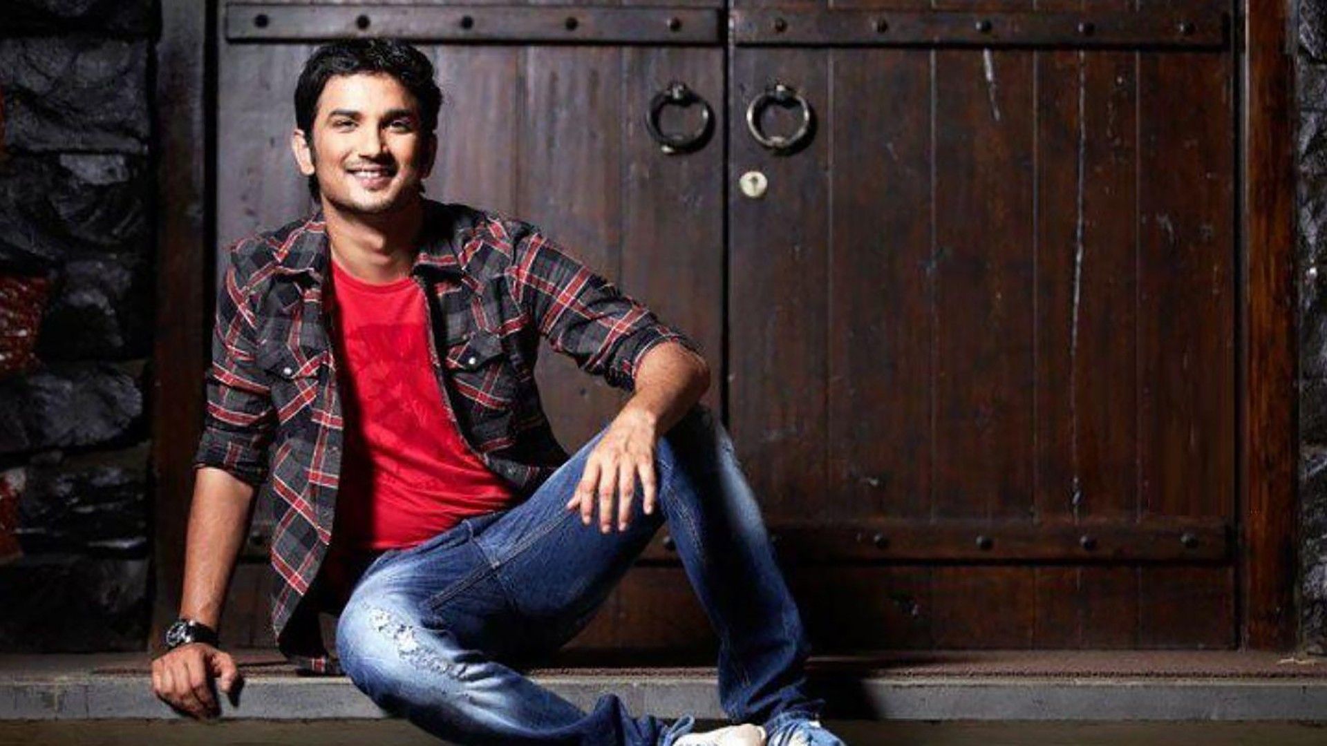 Sushant Is Sitting On Floor Wearing Red Black Shirt And Blue Jean In Wooden Door Background HD Sushant Singh Rajput Wallpaper