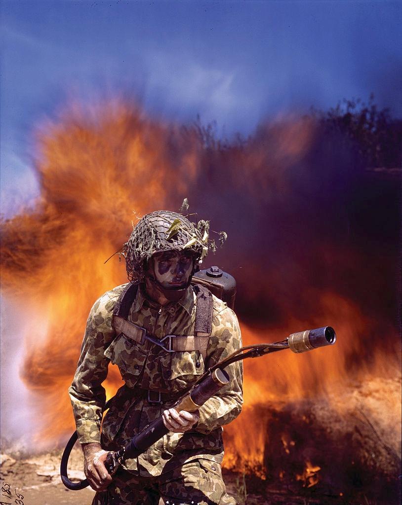 An American soldier dressed in camouflage and face paint holds an M1 flamethrower with fire and smoke behind him during a training exercise in Fort Belvoir, Virginia, c. 1942