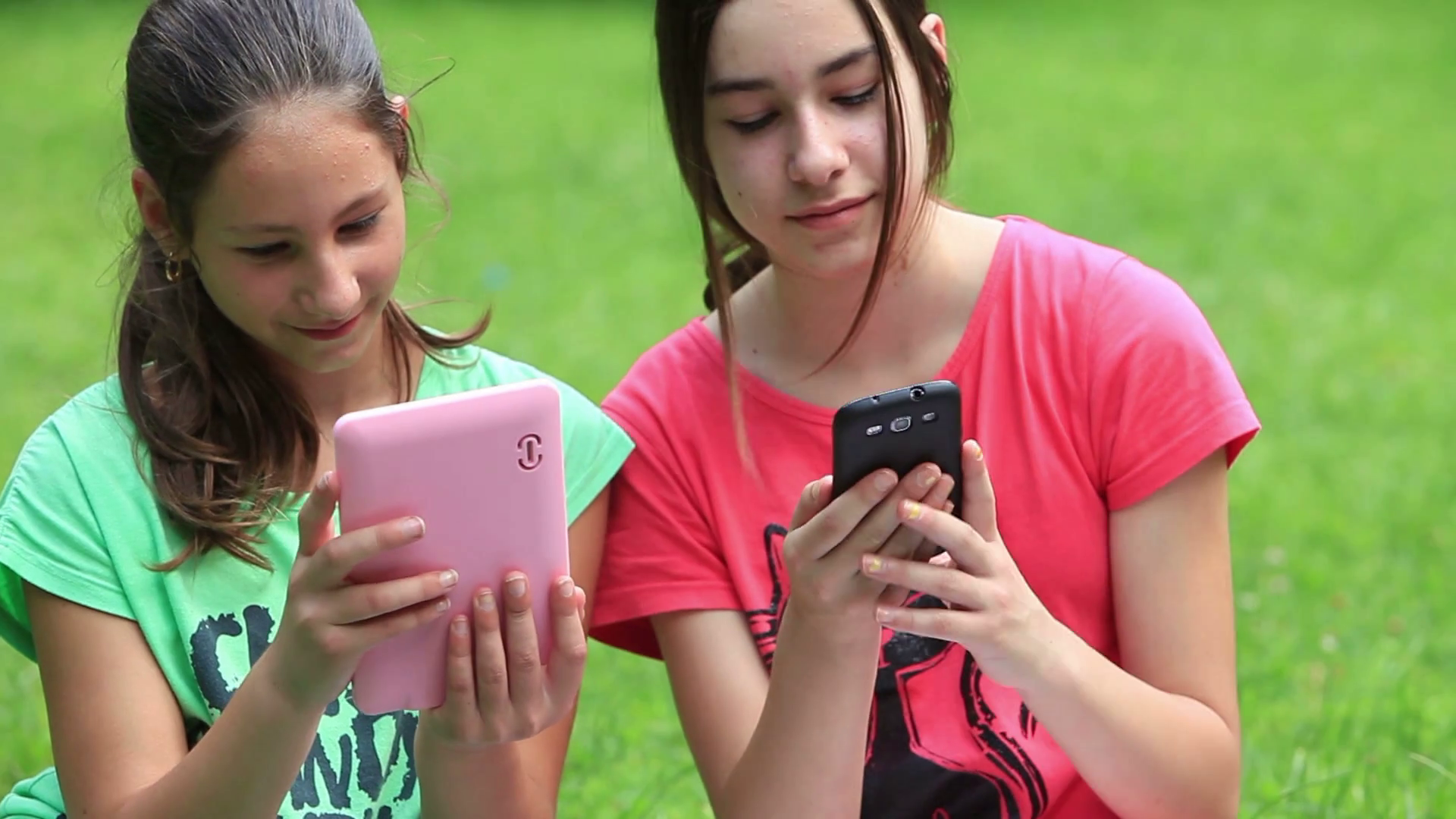 Two girls sitting on a meadow and showing eachother some picture on their devices and giggling. Stock Video Footage