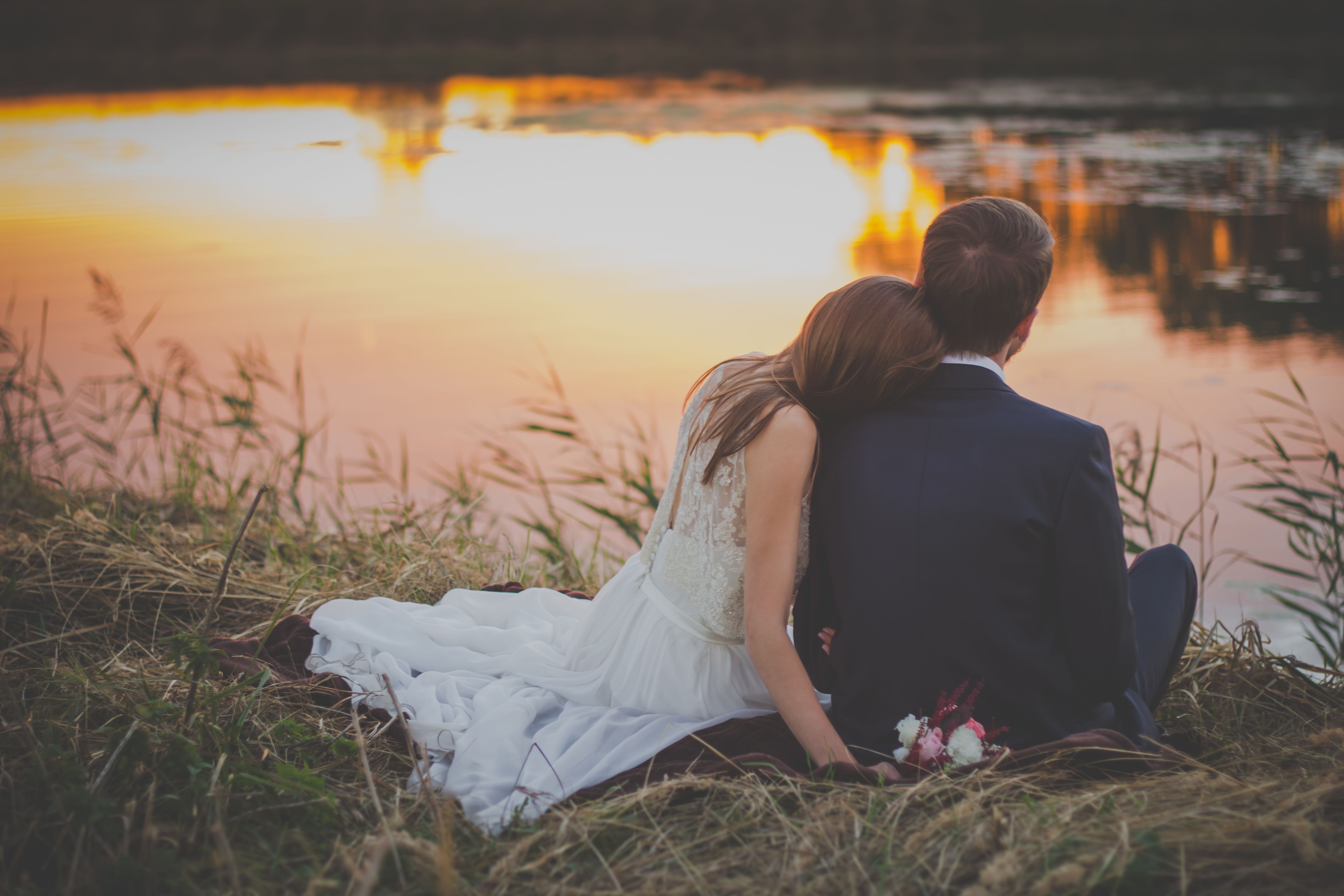 Wedding Couple Sitting on Green Grass in Front of Body of Water at Sunset · Free