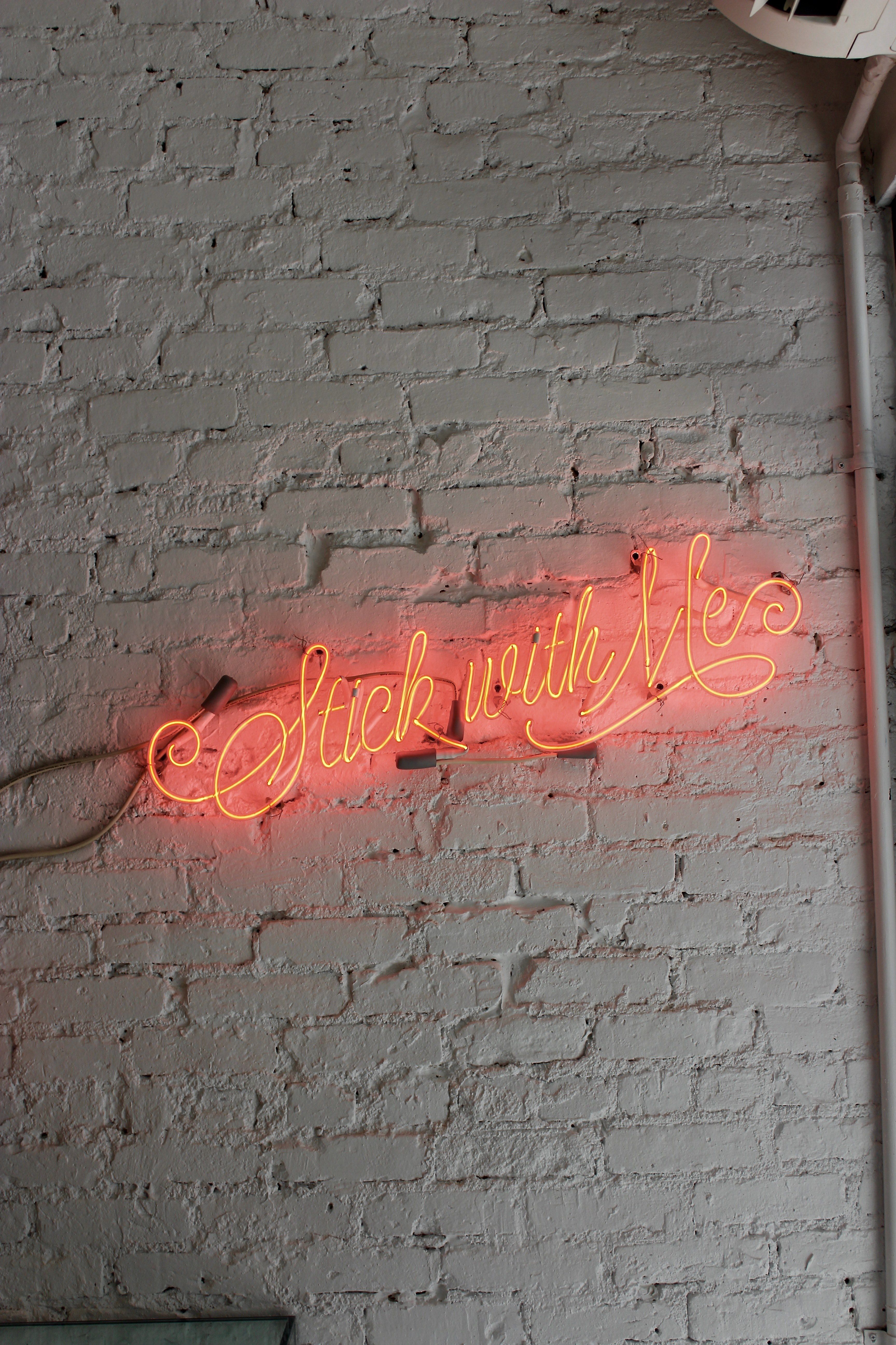 Wallpaper / a red neon with stick with me written in cursive on a white brick wall, stick with me neon 4k wallpaper