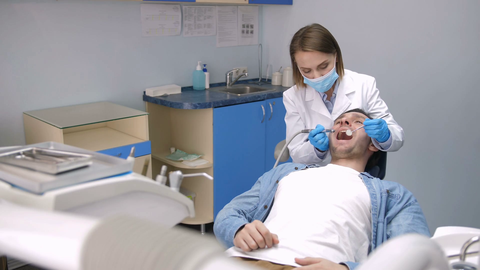 Front view of female dentist in gloves and face mask sitting behind male patient and treating his teeth with drill machine at modern dental clinic. Young patient receiving treatment for cavity Stock