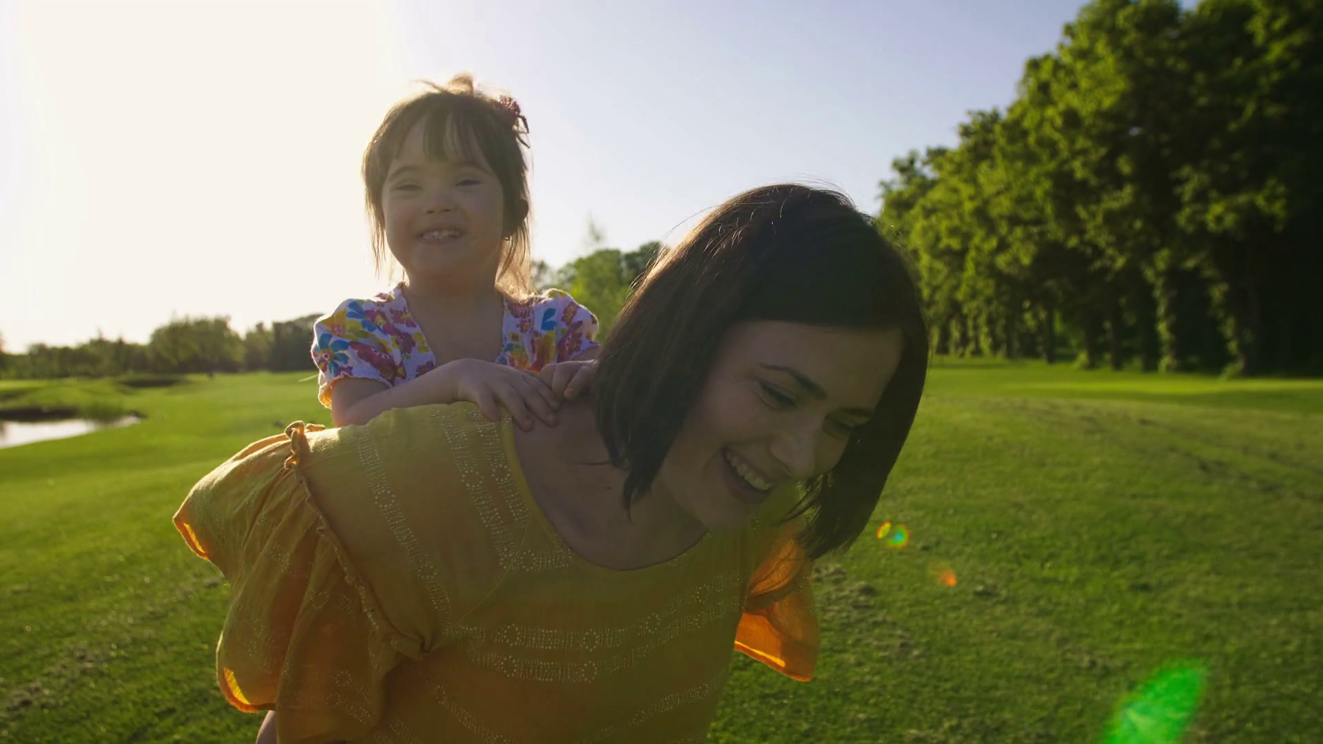 Laughing little toddler girl with down syndrome riding mother's back while mom is running on green grass meadow outdoors in summer. Mom giving piggyback ride to happy daughter in park. Steadicam shot