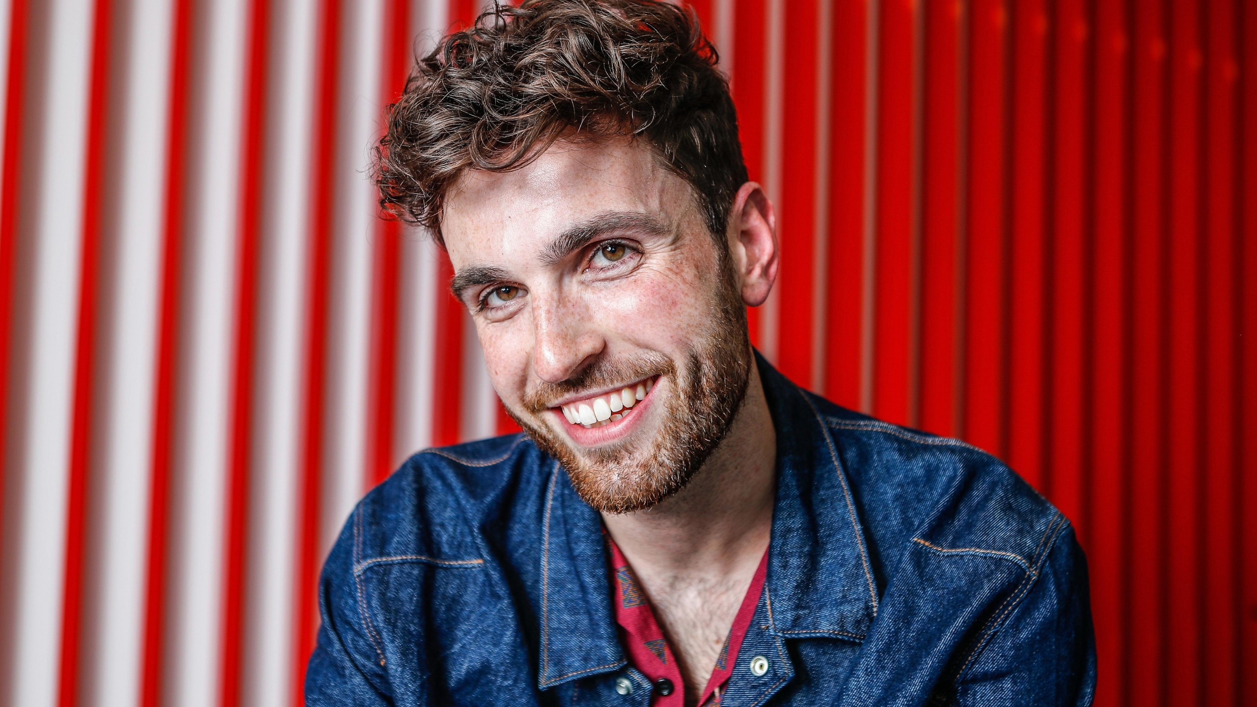 Smiley Duncan Laurence Is Wearing Blue Jean Shirt Sitting In Red Background 4K HD Duncan Laurence Wallpaper